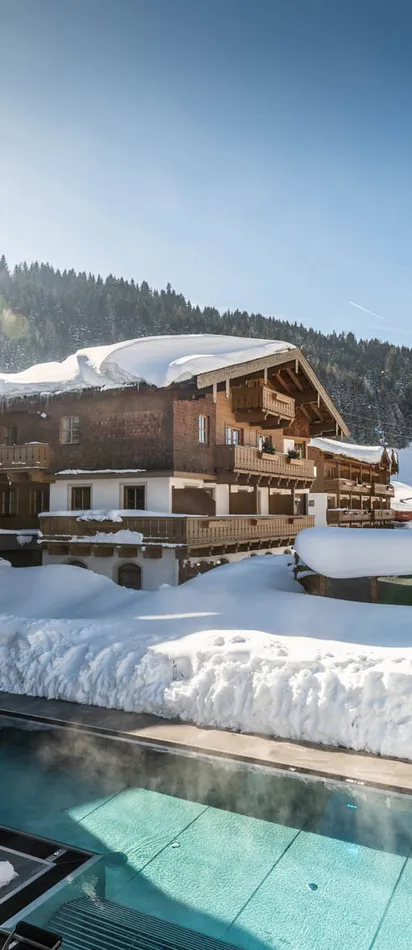 Wellnesshotel mit beheiztem Außenpool in verschneiter Alpenlandschaft bei Sonnenschein