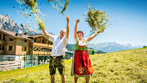 Paar in traditioneller Tracht wirft Heu auf einer Almwiese in den österreichischen Alpen