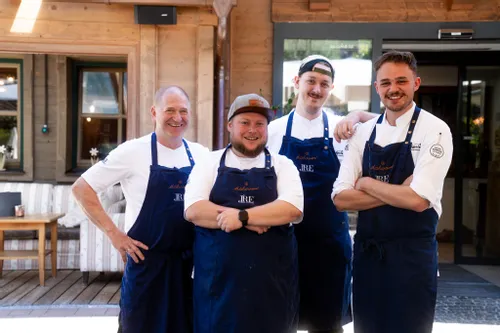 Vier lächelnde Köche in blauen Schürzen vor einem rustikalen Restaurant