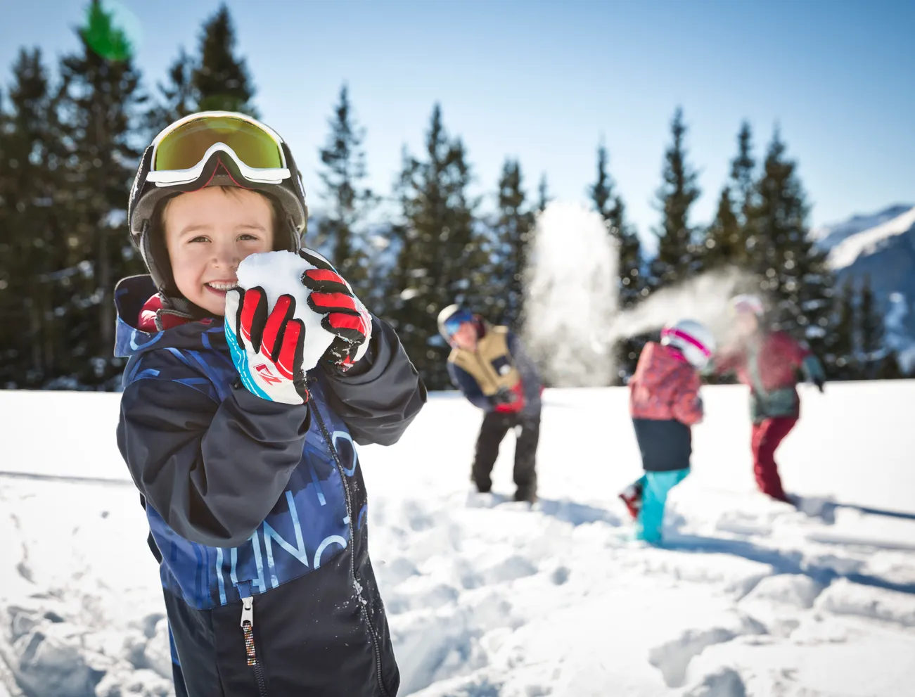 Lächelndes Kind hält Schneeball bei Schneeballschlacht mit anderen Kindern in verschneiter Berglandschaft