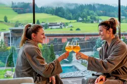 Two women in spa robes toasting drinks with mountain view in wellness resort
