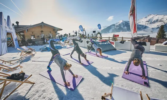 Women doing outdoor yoga on snow in the Alps near a ski lodge under clear blue sky