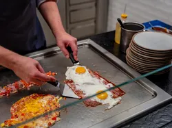 Frühstückszubereitung mit Spiegelei, Speck und Gemüse auf einer heißen Grillplatte