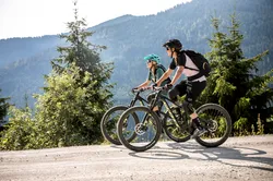 Zwei Mountainbiker fahren bergauf auf einem Waldweg mit alpiner Landschaft im Hintergrund