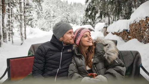 Glückliches Paar in einer Pferdeschlittenfahrt durch verschneiten Wald und Holzstapel