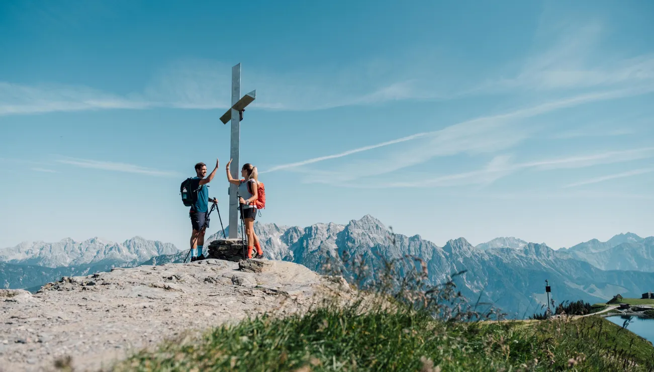 Wanderer geben sich High-Five am Gipfelkreuz mit Alpenpanorama im Hintergrund