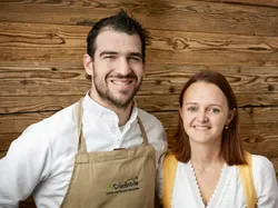 Zwei lächelnde Personen in traditioneller Kleidung vor Holzpaneelwand